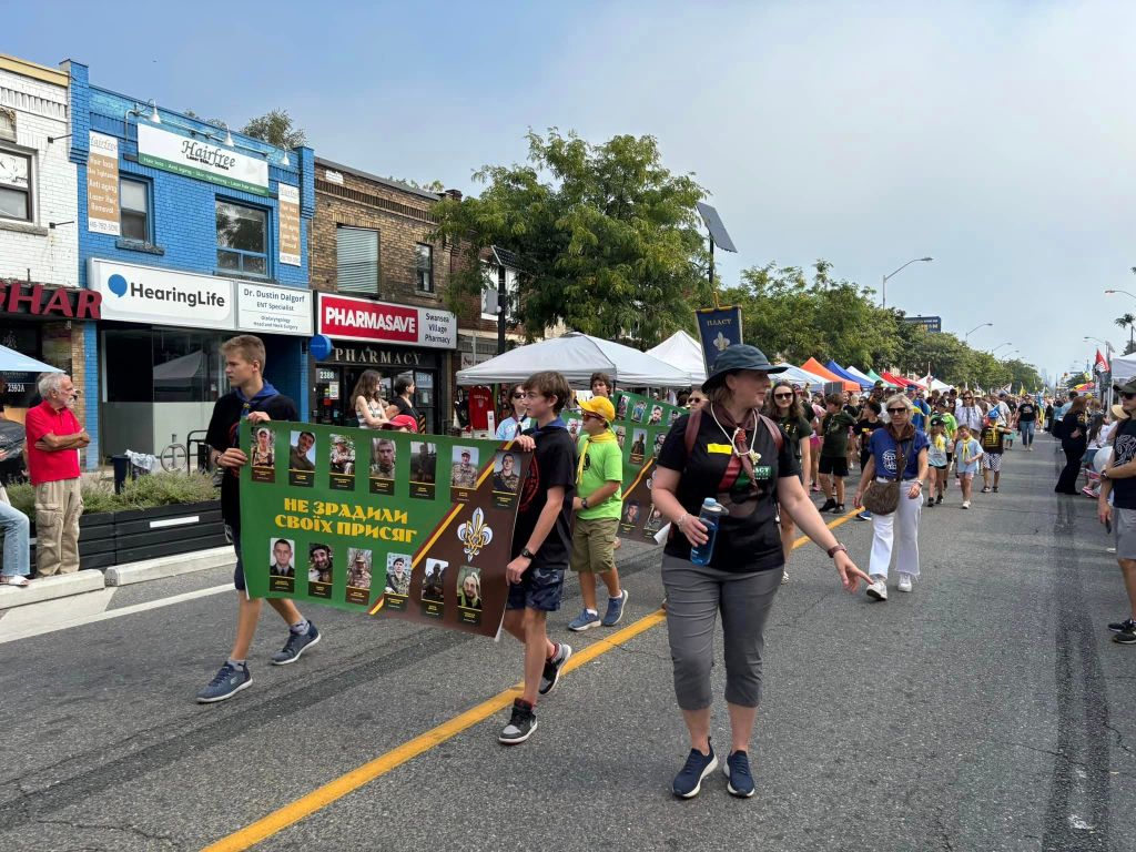 A group of people are participating in a parade, standing on a road and carries Canadian and Ukrainian flags.