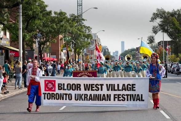 A group of people are participating in a parade, standing on a road and carries Canadian and Ukrainian flags.